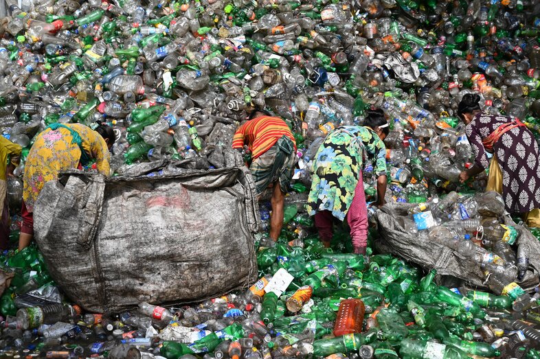 A mountain of plastic bottles towers over workers bent, collecting bottles into large tarp bags at a scrapyard in Bangladesh. The people are dwarfed by the wall of soda and water bottles, shining green and clear in the sun, towering above them.