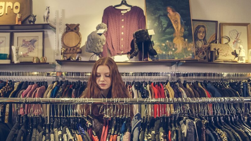 Das Bild zeigt eine Frau, die hinter einer Kleiderstange in einemSecondhand-Laden steht und stöbert