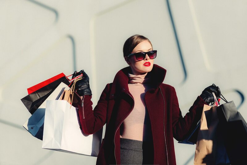 धूप का चश्मा पहने 'स्टाइलिश' महिला सफेद पृष्ठभूमि पर दोनों हाथों में कईशॉपिंग बैग पकड़े हुए है।