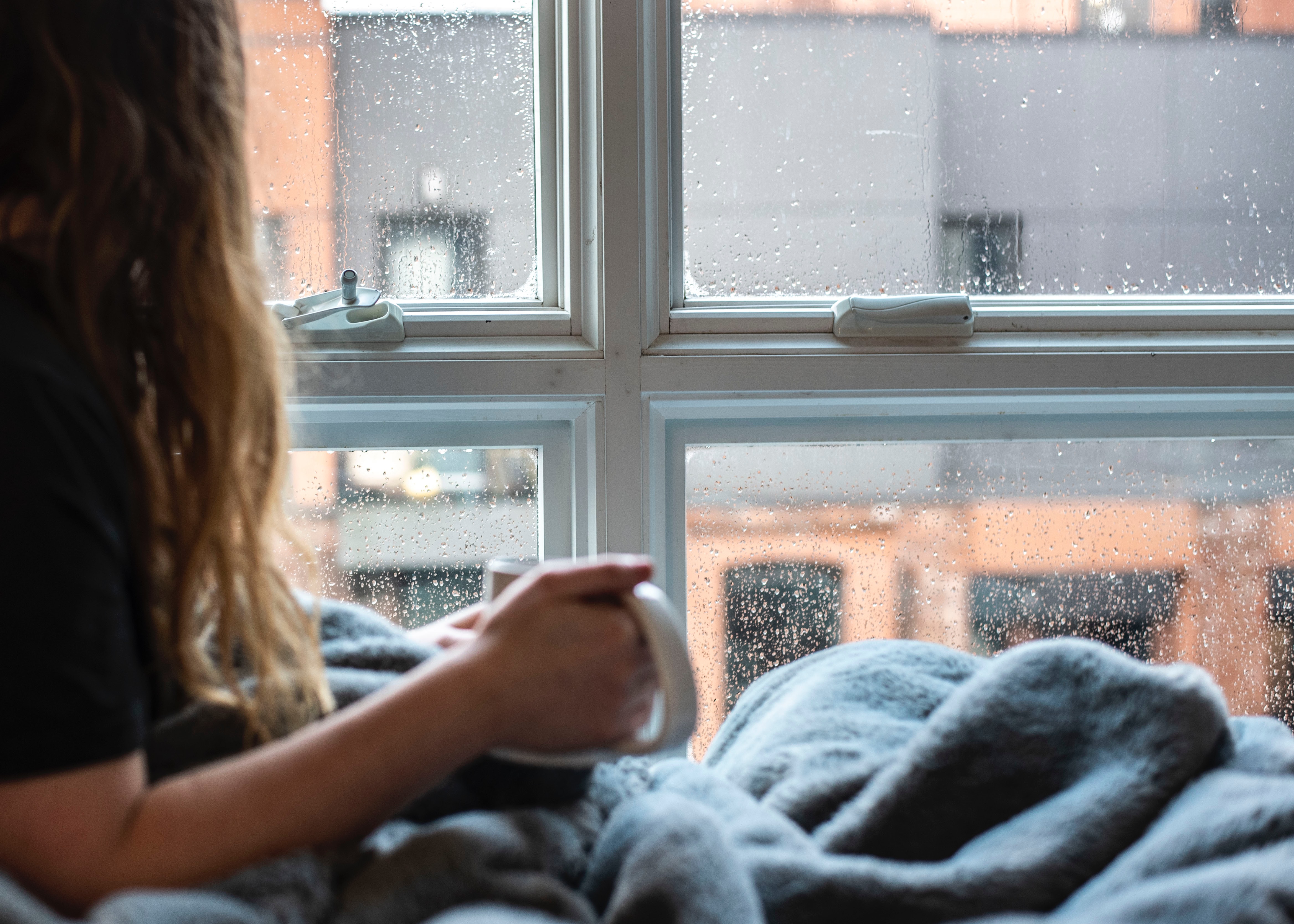 Person seated inside by a rain-spattered window with a blanket and a hotdrink