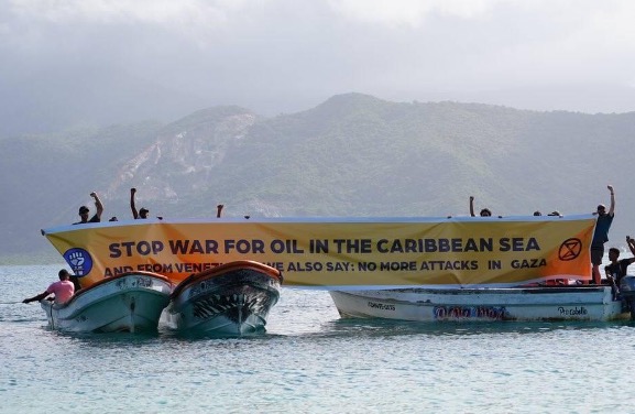 A photograph of three or four small boats in a tight group, the people on the boats holding up a yellow banner that reads "Stop war for oil in the Caribbean Sea".