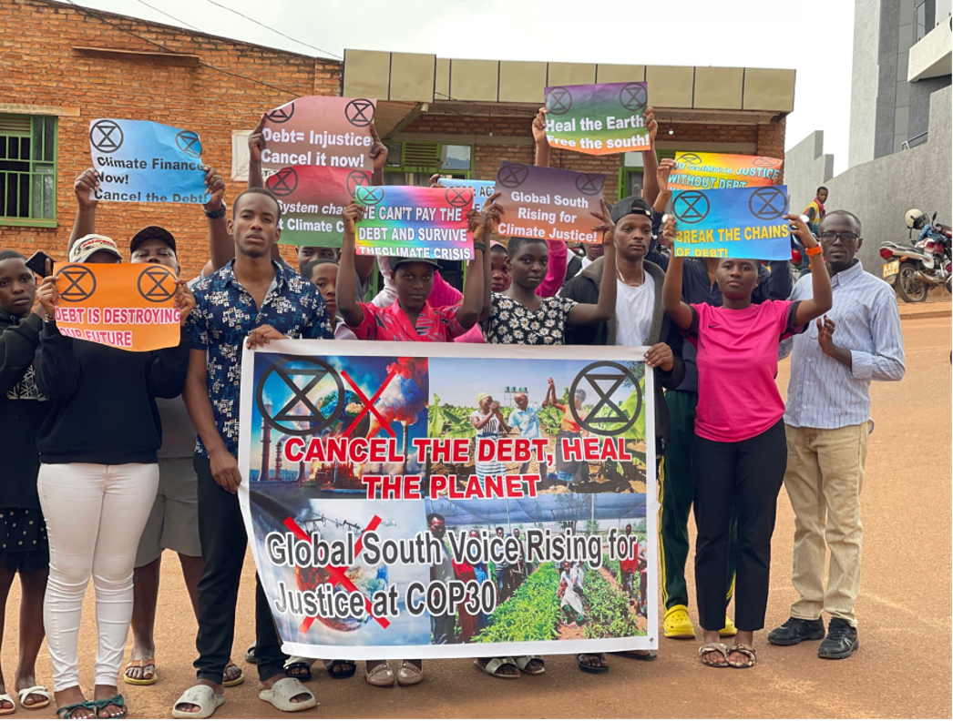 A photograph of a group of people standing in front of a brick building holding signs that bear XR symbols and related slogans. The largest sign reads "cancel the debt heal the planet".