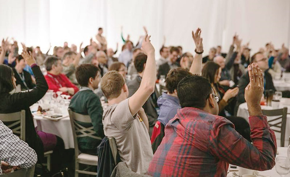 many people seated around tables with their hands up.