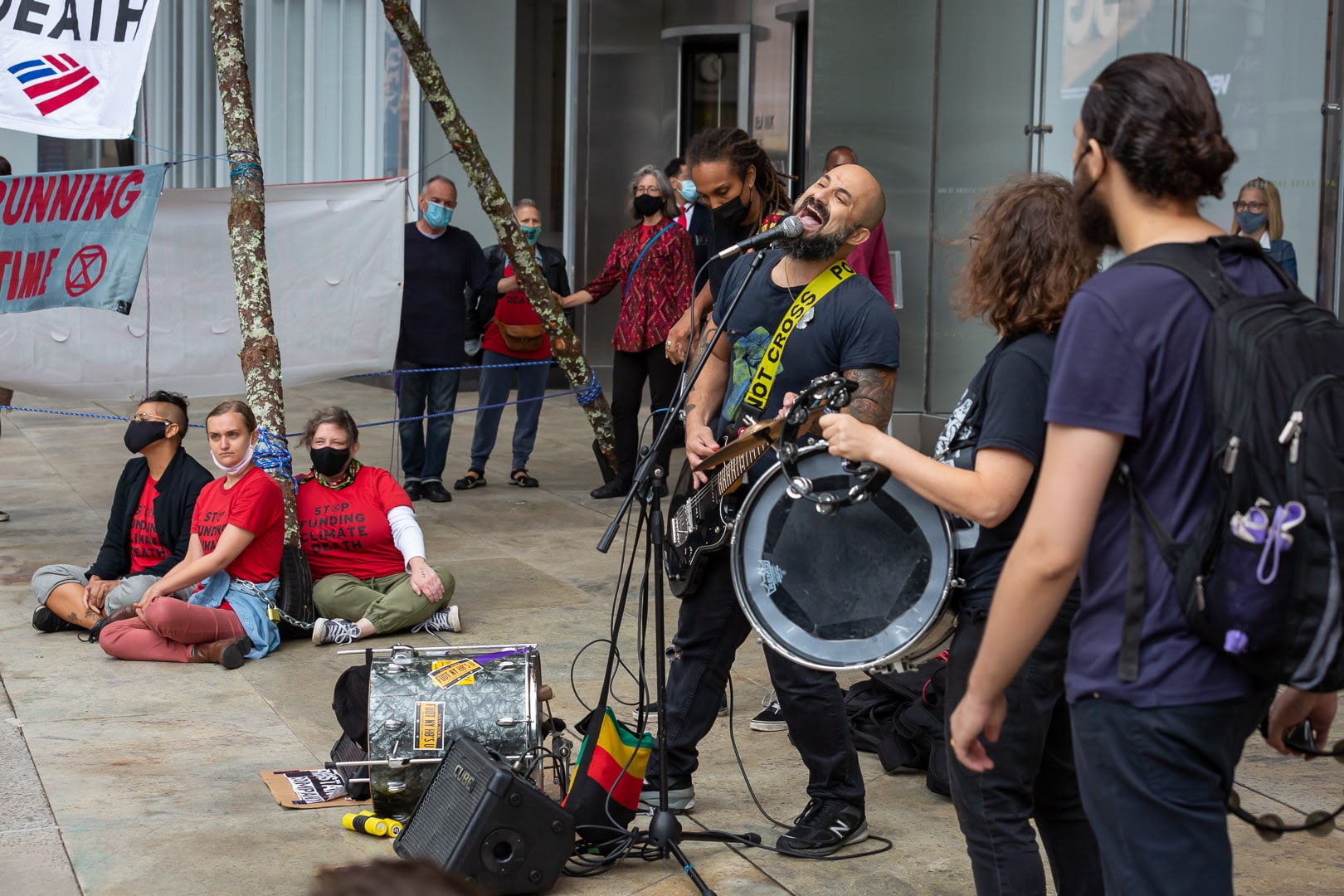 A rock band perform next to a tripod set-up in the forecourt of a skyscraper.