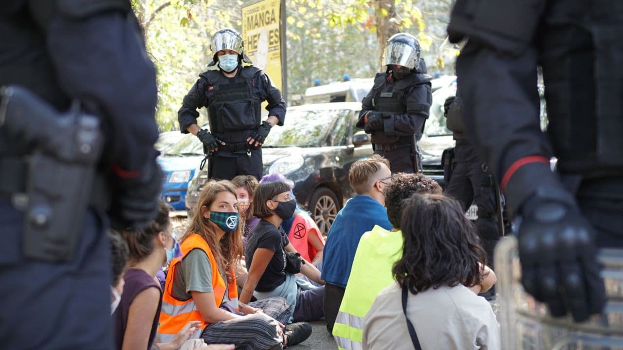 A rebel in an orange high-vis jacket looks at the camera. Seated cross-legged on the ground, she and other rebels are surrounded by officers in riot gear.