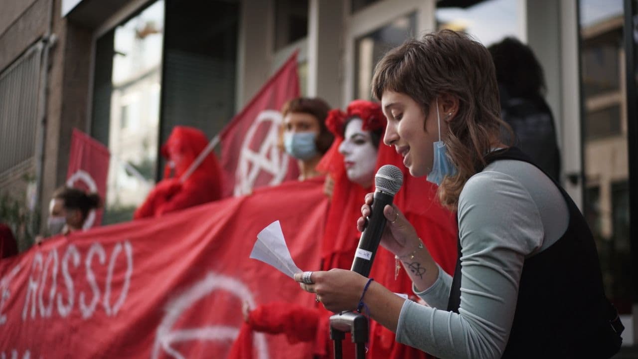 A young rebel speaks into a microphone while other rebels hold up a red banner outside the entrance to Rai's HQ.