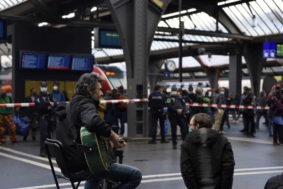 A man sits in the foreground with a guitar surrounded by a police barricade and officers in the middle of a train station.