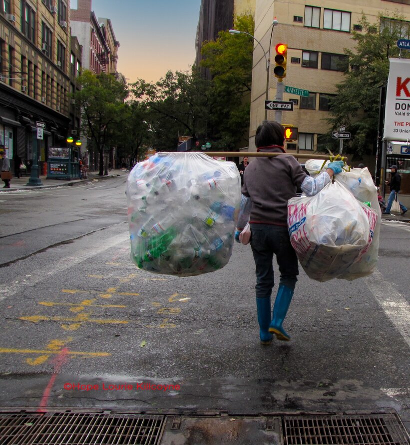 A person crosses the street in New York City as evening falls. They carry a wooden pole across their right shoulder, with two huge translucent white plastic bags balancing on each end, bulging with empty plastic bottles.