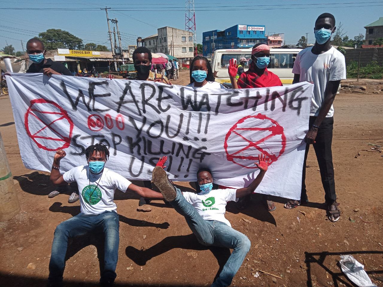 A group of XR Kisumu rebels hold a sign reading “We are watching you!!! Stop killing us!!!” Two rebels lie on the ground in front of the sign, one with his hands painted red.