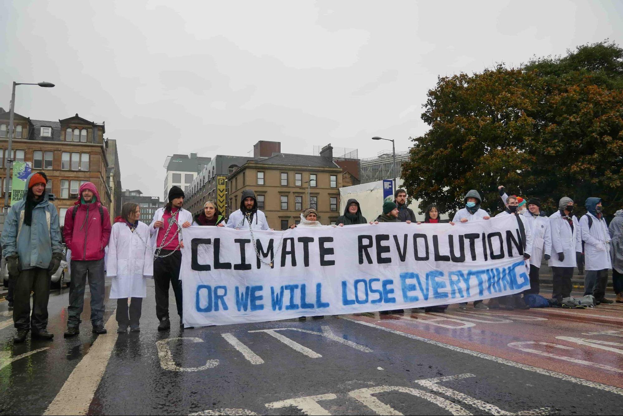 Scientists dressed in white coats stand in a long line across a road holding a sign which says 'climate Revolution or we will lose EVERYTHING'