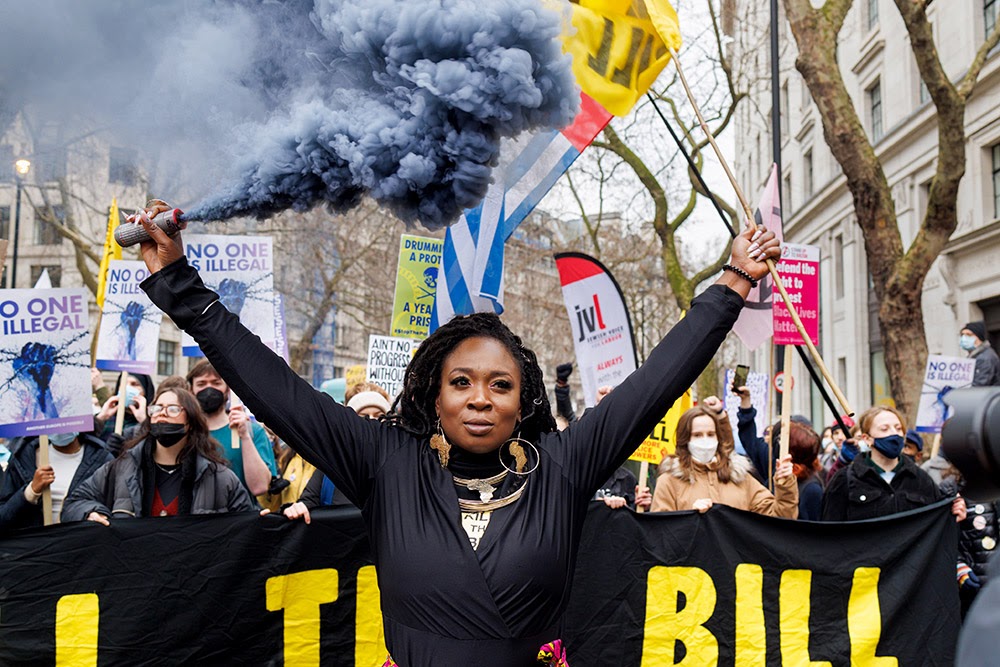 A woman holds a flare and a 'Kill The Bill' flag. She is standing in front of a protest march, at the front of which protestors are holding a long black 'Kill The Bill' banner. 