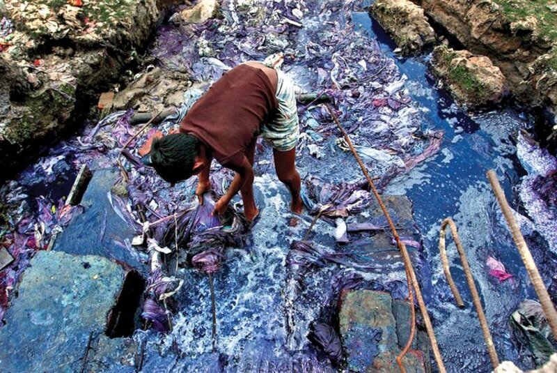 Image shows boy washing clothes in river stained with dyes, view from above.