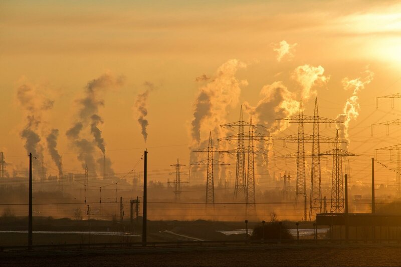 Image shows a line of smoke plumes from factories set against a yellowsunset with powerlines in theforeground.