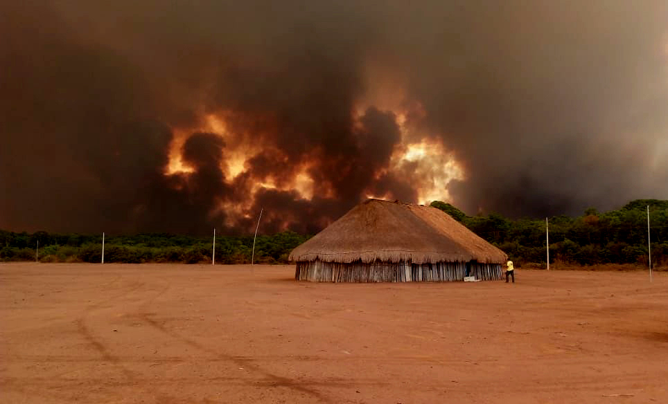 A wooden house sits on an arid plain with a great fire burning in the background.