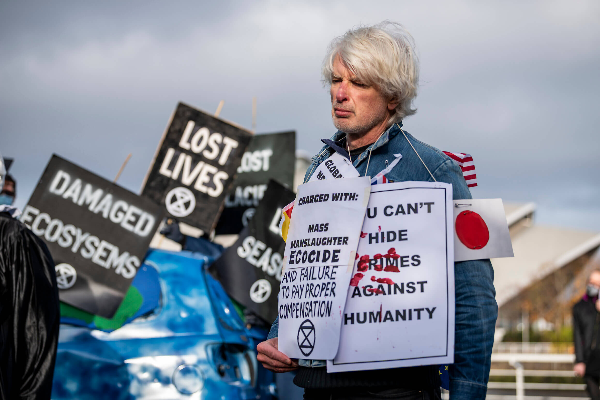 Image shows an XR activist wearing signs saying "Charged with Mass Slaughter, Ecocide and Failure to Pay Proper Compensation"