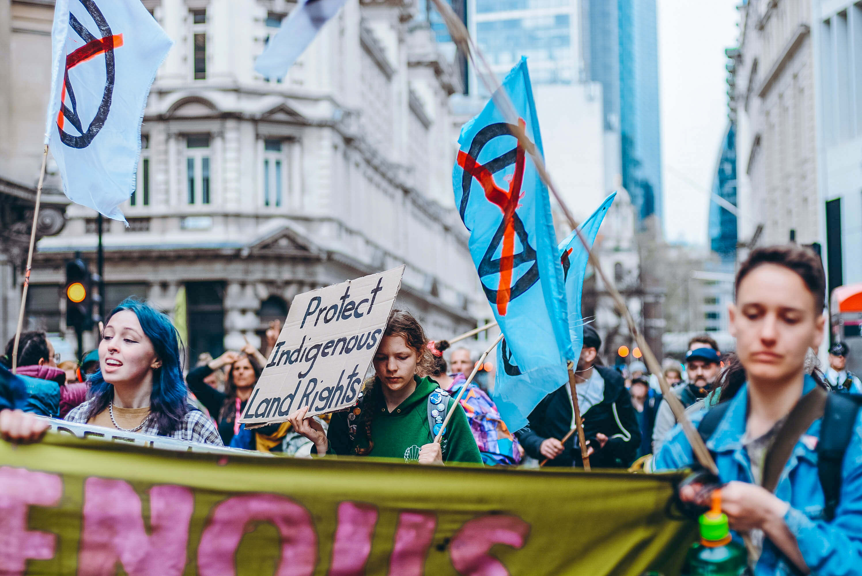 Image shows XR activists on the streets of Glasgow during COP26, holding a banner, a sign saying "Protect Indigenous Land Rights" and waving flags