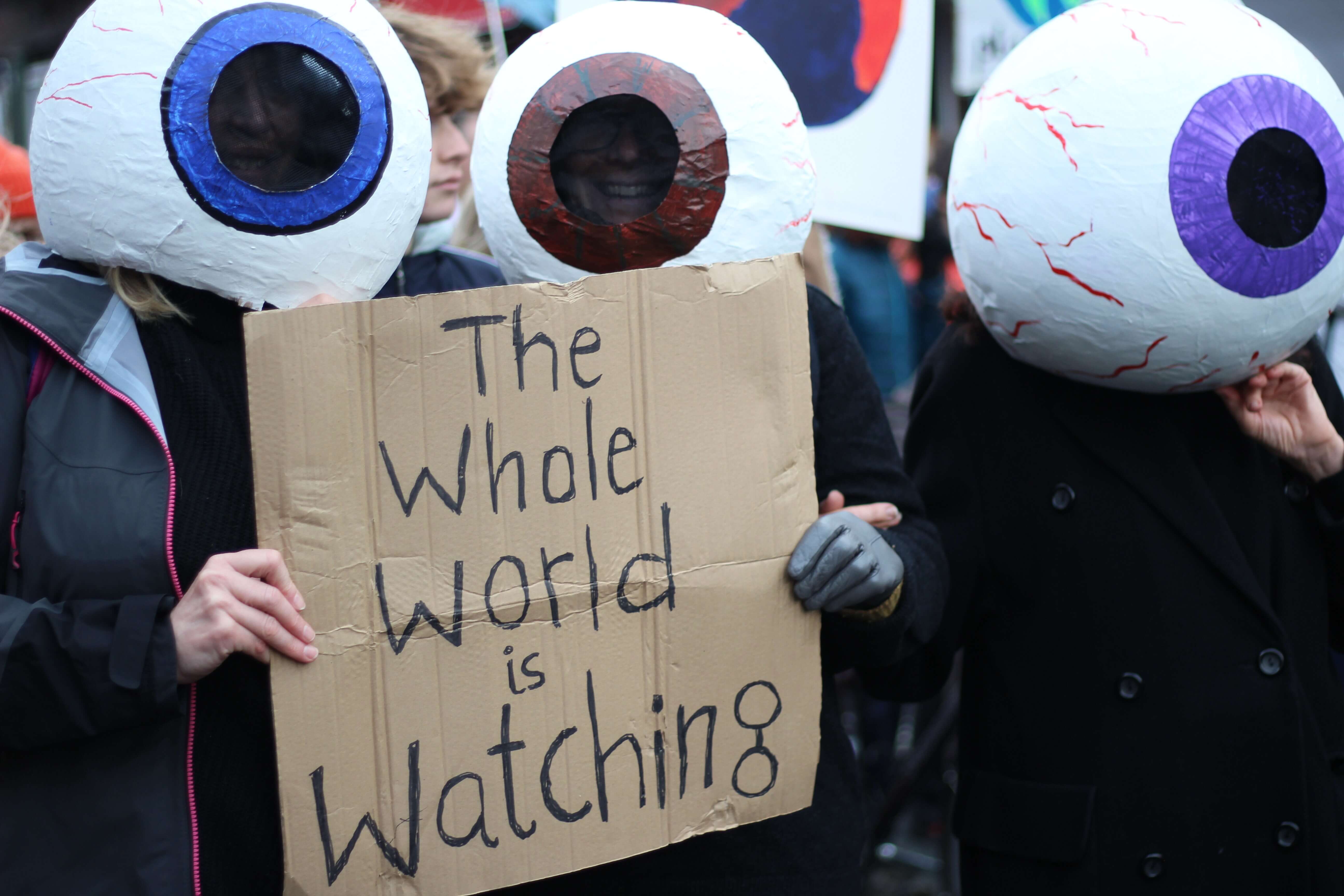 Image shows activists with papier mache eyes on their heads holding up a sign saying "The Whole World is Watching"
